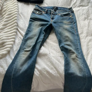 True Religion Joey Low Rise Flare Jeans - Blå jeans från True Religion, modell Joey Low Rise Flare. Extremt slitna med utställda ben och låg midja. Klassiska orange sömmar och detaljerade fickor. Osäker på insidan men är där för att hålla ihop byxorna pga tidigare hål. Användna för mycket. Finns också fläck på framsidan, kan skicka mer bild.