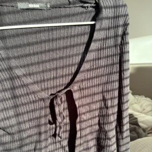 Svart ribbad kofta från BikBok - Svart ribbad bolerotopp från BikBok med lång ärm och croppad passform. Toppen har v-ringning och knyt framtill, vilket ger en trendig och avslappnad look. Perfekt att styla över ett linne eller klänning för en extra touch.