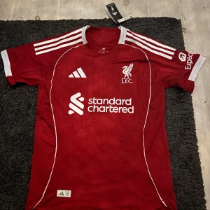 Liverpool FC matchtröja Adidas röd M - Liverpool FCs officiella matchtröja från Adidas i rött med vita detaljer och klubbmärke på bröstet. Slim fit-modell med korta ärmar, klassiska Adidas-ränder på axlarna och Standard Chartered-tryck framtill. Tillverkad i lätt och ventilerande funktionsmaterial.
