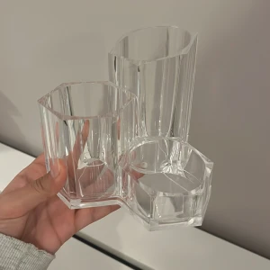 Organisation  - Denna produkt är en transparent, hexagonformad plastbehållare med tre fack, perfekt för förvaring av pennor, sminkborstar eller småsaker. Den är i gott skick utan synliga repor eller skador. Modern och stilren design som passar på skrivbord eller i badrum.