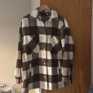 Brun och vit rutig skjorta från Divided - Säljer en oversized skjorta/kan användas som jacka, brun och vit rutig overshirt från Divided. Jackan har stora bröstfickor, knappar framtill och klassisk krage. Perfekt lager-på-lager-plagg med lång ärm och mjukt material som känns skönt mot huden.