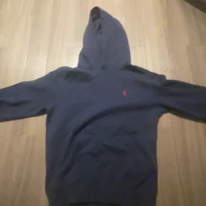 Mörkblå hoodie från Polo Ralph Lauren - Snygg mörkblå hoodie från Polo Ralph Lauren med klassisk röd logga broderad på bröstet. Tröjan har huva, långa ärmar och är tillverkad i en mjuk bomulls- och polyestermix. Perfekt för chill dagar eller när du vill ha en stilren streetlook. OBS!! Snöre saknas=varför priset är 1200kr.