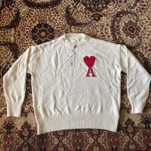 Vit stickad tröja från Ami Paris - Vit stickad tröja från Ami Paris med rund halsringning och ribbade muddar. På bröstet finns ett stort rött hjärta med bokstaven A. Tröjan är i bomull och har en clean, stilren look som sticker ut med den ikoniska loggan.