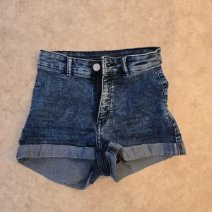 Blå högmidjade jeansshorts - Snygga blå jeansshorts med hög midja och uppvikta benslut. Klassisk femficksmodell med bälteshällor och knapp- samt dragkedjestängning. Perfekta för varma dagar och enkla att matcha med allt. Tidlös denim-look som alltid funkar.