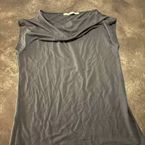 Mörkblå ärmlös topp med drapering - Snygg mörkgrå ärmlös topp med draperad halsringning. Enkel och stilren design som passar perfekt till jeans eller kjol. Mjuk och skön känsla i materialet. 