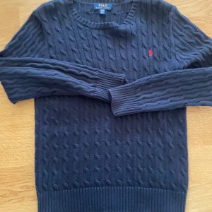 Mörkblå kabelstickad tröja Polo Ralph Lauren - Kabelstickad mörkblå tröja från Polo Ralph Lauren med klassisk rund halsringning och röd broderad logga på bröstet. Långärmad modell i mjuk bomull, perfekt för lager-på-lager och snygg till jeans.