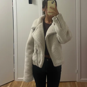 Off-white teddyjacka med dragkedja - Säljer en off-white teddyjacka med oversized krage och cool dragkedjedetalj. Jackan är fluffig och har en loose passform som ger en chill vibe. Perfekt för dig som vill ha en mysig och trendig look under höst och vinter.