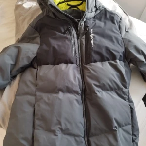 Grå pufferjacka från Sail Racing - Säljer en grå pufferjacka från Sail Racing med svart ovandel och huva. Jackan har två bröstfickor med dragkedja och en hel dragkedja framtill. Insidan är fodrad i neonfärg. Perfekt för kalla dagar och riktigt snygg streetstil.