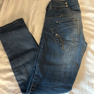 Blå jeans från Sublevel med detaljer - Snygga blå jeans från Sublevel med flera knappar i midjan och unika sömdetaljer vid fickorna. Jeansen har en klassisk femficksdesign och är tillverkade i slitstarkt denim. Perfekta för dig som gillar en cool och avslappnad stil.