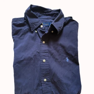 Marinblå kortärmad skjorta Ralph Lauren - Snygg marinblå skjorta från Ralph Lauren med klassisk krage och korta ärmar. Skjortan har knappar framtill och den ikoniska broderade loggan på bröstet. Tillverkad i mjuk bomull som känns skön mot huden. Perfekt för dig som gillar stilrena och tidlösa plagg.