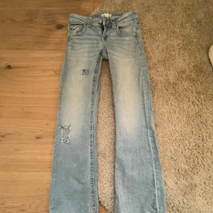 Ljusblå s jeans Gina Tricot med hål - Ljusblå jeans från Gina Tricot i storlek 152. Jeansen har slitna detaljer på benen och klassisk femficksmodell. De är raka i modellen och har dragkedja och knapp framtill. Perfekta för en avslappnad och trendig look.