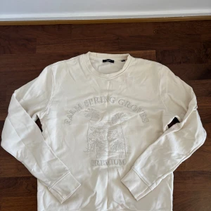 Vit sweatshirt från Jack & Jones - Säljer en stilren vit sweatshirt från Jack & Jones med broderad text och palm-motiv framtill. Tröjan har rund hals, långa ärmar och är tillverkad i mjuk bomull.  Lite skrynklig så inte använt på länge. 