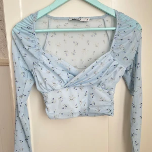 Blommig omlottopp från NA-KD ljusblå - Supersöt blommig ljusblå omlottopp från NA-KD. Perfekt för dig som gillar romantisk och feminin stil. Endast använd fåtal gånger. 