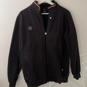 Svart Puma AMG ziptröja - Svart långärmad ziptröja från Puma x Mercedes AMG med broderad Mercedes-logga fram och stor AMG-tryck på ryggen. Hög krage med röd och gul rand, två dragkedjor framtill och ribbade muddar. Perfekt för dig som gillar motorsport och streetwear.