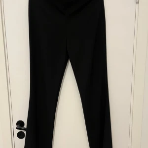 Svarta kostymbyxor med bootcut - Svarta kostymbyxor från C i storlek 38. Byxorna har en klassisk bootcut-modell med hög midja och diskret dragkedja i sidan. Perfekta för en clean och stilren look. Materialet känns mjukt och följsamt.