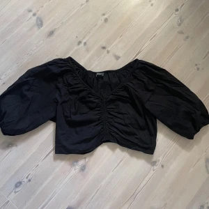 Svart croppad blus från Gina Tricot - Säljer en svart croppad blus från Gina Tricot med v-ringning och puffiga trekvartsärmar. Toppen har rynkade detaljer framtill och resår vid halsringning och ärmslut. Perfekt att styla med höga jeans eller kjol.