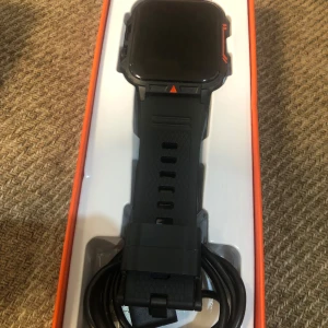 Svart sportig smartwatch med laddare - Säljer en svart Amazfit smartwatch med robust, fyrkantig design och silikonarmband med mönstrad yta. Klockan har orange detaljer runt boetten och kommer med laddningskabel. Perfekt för dig som gillar teknik och vill ha koll på träningen.