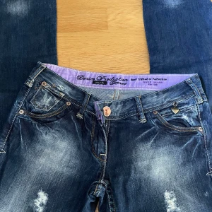 River Island blå jeans med hjärtdetalj - Snygga blå jeans från River Island med slitna detaljer och dekorativt hjärthänge vid fickan. Jeansen har klassisk femficksdesign, kontrastsömmar och coola bakfickor med zigzag-mönster. Perfekt för dig som gillar en trendig och avslappnad look.
