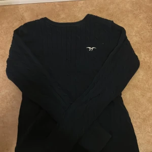 Mörkblå stickad tröja Hampton Republic - Snygg mörkblå stickad tröja från Hampton Republic med diskret logga på bröstet. Tröjan har klassiskt kabelstickat mönster och långa ärmar. Perfekt för dig som gillar preppy stil och vill ha något som funkar året runt.