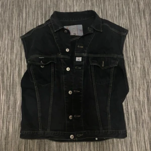 Svart jeansväst från Fashion Victim - Svart jeansväst från Fashion Victim med klassisk krage, kontrastsömmar och knappar framtill. Västen har två bröstfickor med lock och knapp samt coola detaljer som ger en edgy look. Perfekt att slänga över en hoodie eller t-shirt. Frakt ingår ej💕