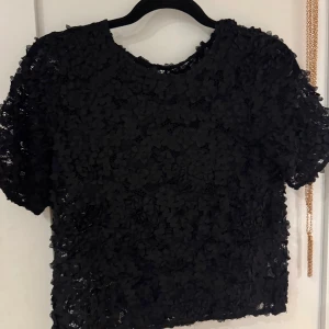 Svart blommig spetsblus från Stockh LM - Supersnygg svart blus från Stockh LM med korta ärmar och täckt av 3D-blommor i spets. Blusen har rund halsringning och är genombruten med ett unikt blommigt mönster som ger en lyxig känsla. Perfekt för dig som vill sticka ut med en cool och elegant topp.