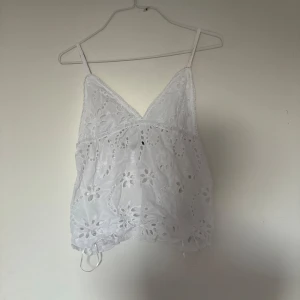 Vit broderad linnetopp med spets - Söt vit linnetopp med tunna axelband och v-ringning. Toppen har broderade blommor och spetsdetaljer, samt en luftig och lätt känsla. Perfekt för varma dagar och ger en romantisk vibe till din outfit.