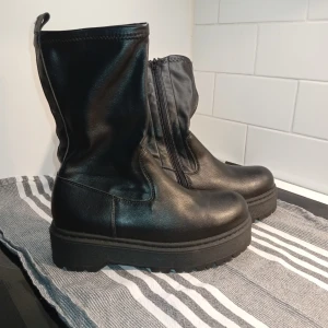Svarta boots med platåsula och dragkedja - Säljer ett par svarta boots i skinnimitation med chunky platåsula och grovt mönstrad undersida. Skorna har dragkedja på insidan och en högre skaftmodell som ger en cool och edgy vibe. Perfekta till höstens outfits och funkar lika bra till jeans som kjol.
