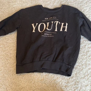 Svart sweatshirt från Kappahl YOUTH - Cool svart sweatshirt från Kappahl med trycket 'YOUTH' och 'New York City' framtill. Tröjan har rund hals, ribbade muddar och är långärmad. Perfekt för dig som gillar en avslappnad och trendig stil.