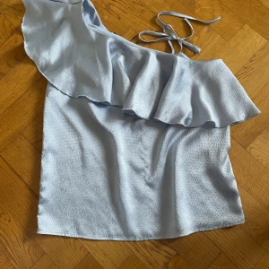 Ljusblå omlottopp från Gina Tricot - Supersöt ljusblå omlottopp från Gina Tricot med volangdetalj längs ena axeln och knytband. Toppen är ärmlös och har en lätt glansig, strukturerad yta som ger en lyxig känsla. Perfekt för varma dagar och snygg till både jeans och kjol.