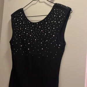 Svart klänning med pärldetaljer - Svart ärmlös klänning med rund halsringning och glittrande pärlor över hela överdelen. Snygg och stilren design som ger en lyxig känsla. Perfekt för dig som vill sticka ut med elegans och coola detaljer.