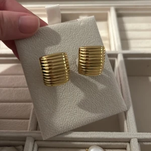Guldpläterade ribbade örhängen muli collection - Snygga örhängen i guldpläterad metall med ribbad från muli collection, rektangulär form. De har en chunky och modern look som verkligen sticker ut. Perfekta för dig som gillar statement-smycken med en cool och stilren vibe. Ordinarie 599kr, pris 300kr