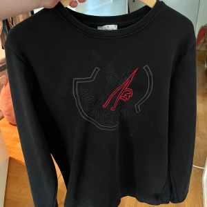 Svart sweatshirt från Moncler XL barn  - Svart sweatshirt från Moncler i storlek XL barn med stort broderat Moncler-logo på bröstet i svart och rött. Tröjan har rund halsringning och långa ärmar, perfekt för en clean och stilren look. Sitter som S i vuxen storlek 