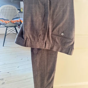 Rutig grå kostymbyxa från Hackett - Snygga grå kostymbyxor från Hackett London i klassiskt rutigt mönster. Byxorna har raka ben, bälteshällor och bakficka med knapp. Tillverkade i en exklusiv ull- och silkesblandning för en lyxig känsla. Perfekta för en dressad look. Passform är åt det smalare hållet.