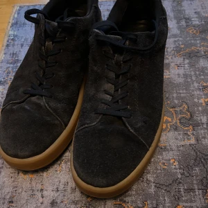 Vagabond Skor med brun sula  - Snygga svarta sneakers i mocka med klassisk rund tå och svarta snören. Skorna har en stilren design med kontrasterande brun gummisula som ger en cool touch. Perfekta för dig som gillar minimalistisk stil och vill ha ett par schyssta vardagssneakers. Okej skick