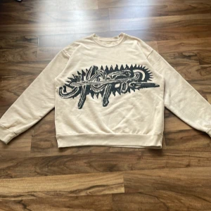 Travis Scott Utopia merch - Säljer en beige UTOPIA crewneck i storlek L. Tröjan har ett stort svart grafiskt tryck framtill och är tillverkad i mjuk bomullsmix med borstad insida. Klassisk passform med ribbade muddar vid ärmslut och midja. Perfekt för dig som gillar streetwear och unika prints. DM om ni också vill ha lådan, den kostar dok extra 
