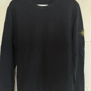 Svart långärmad tröja Stone Island - Svart långärmad tröja från Stone Island med klassisk rund hals och ikonisk logotyp-patch på vänster ärm. Tröjan är i mjuk bomull och har en clean, stilren look. 