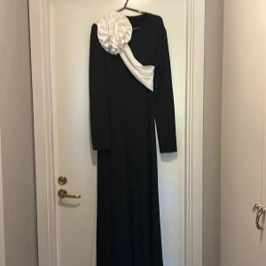 Svart långklänning med vit rosett SHEIN - Svart långklänning från SHEIN med lång ärm och en stor vit, glansig rosett i satin över axeln som fortsätter ner längs sidan. Klänningen är figurnära och har en unik design med kontrasterande detaljer. Materialet är mjukt och stretchigt i polyester och elastan.