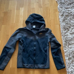Svart och grå Nike hoodie XS - Nike hoodie i svart och grått med guldig logga på bröstet. Tröjan har dragkedja hela vägen, huva och långa ärmar. Perfekt för chill eller träning, med en snygg och sportig look.