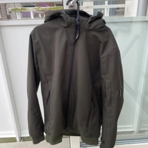 Grön softshell/vindjacka från C.P. Company - Snygg mörkgrön vindjacka från C.P. Company med huva och ikonisk linsdetalj på ärmen. Jackan har dragkedja framtill, två fickor med dragkedja och ribbade muddar. Perfekt för dig som gillar streetwear och stilren design. Prislapp ligger vid sidan av och är placerad i jackans ficka vid försäljning.