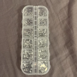 Strass-stenar för smink och naglar - Ett set med små, runda strass-stenar i olika storlekar, perfekta för att dekorera sminklooker eller naglar. Stenarna är silvriga och kommer i en genomskinlig plastask med flera fack för enkel sortering.