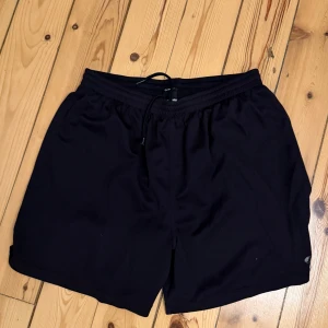 Svarta träningsshorts från SOC - Svarta träningsshorts från SOC i storlek L. De har elastisk midja med snörning och är tillverkade i 100% polyester för en lätt och luftig känsla. Perfekta för gymmet eller löprundan, med klassisk och enkel design.