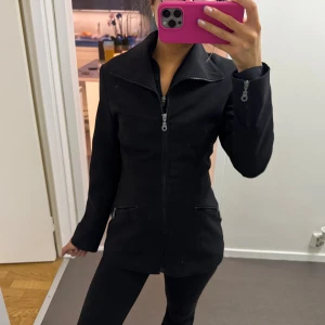 Svart kavaj med dragkedja - Snygg svart kavaj med markerad krage och hel dragkedja framtill. Jackan har två fickor med dragkedja och långärmad design. Passformen är slim , perfekt för en clean och stilren look.