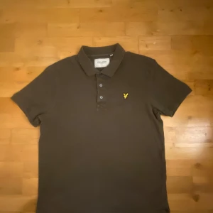 Brun pikétröja från Lyle & Scott - Snygg brun pikétröja från Lyle & Scott med klassisk krage och tre knappar framtill. Tröjan har det ikoniska gula fågel-loggan broderad på bröstet och är tillverkad i bomull. Perfekt för en clean och stilren look. Size L och är i mycket fin skick. Om det finns några frågor så är det bara att fråga!