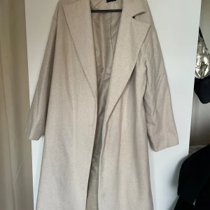 Beige kappa från Boohoo - Säljer en snygg beige kappa från Boohoo i storlek 38/40. Jackan har klassisk krage, lång ärm och ett diskret fiskbensmönster. 