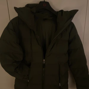 Mörkgrön pufferjacka från Bergans - Säljer en mörkgrön pufferjacka från Bergans i storlek S. Jackan har hög krage, huva och två fickor med dragkedja framtill. Perfekt för kalla dagar och har en clean, stilren look med diskret logga på bröstet.