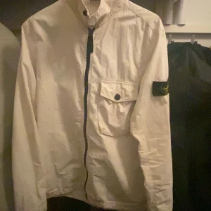 Vit overshirt från Stone Island - Säljer en clean vit overshirt från Stone Island med klassisk patch på ärmen. Jackan har dragkedja framtill, bröstficka med knapp och en avslappnad passform. Finns en liten fläck (se sista sliden) men inget man märker. Priset kan diskuteras vid snabb affär.