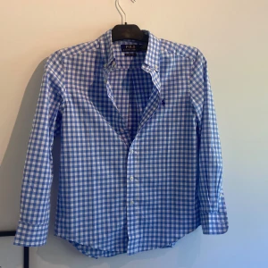 Blårutig skjorta från Polo Ralph Lauren - Snygg blårutig skjorta från Polo Ralph Lauren i bomull med naturlig stretch. Skjortan har klassisk krage, knappar framtill och långa ärmar. Perfekt för dig som gillar stilrena och fräscha plagg med en preppy vibe. Borde passa dig runt 160 bäst!