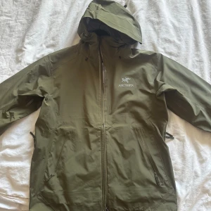 Olivgrön regnjacka från Arc'teryx - Säljer en olivgrön regnjacka från Arc'teryx med huva och hel dragkedja framtill. Jackan har två snedställda fickor med dragkedja och justerbar huva. Perfekt för blöta dagar och outdoor-äventyr. Snygg minimalistisk design med Arc'teryx-logga på bröstet.