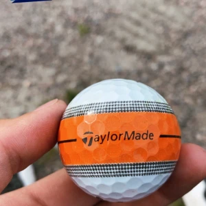 TaylorMade Golfboll - TaylorMade golfboll med orange och svart design. Bollen är i  gott skick utan synliga repor eller skador. Perfekt för golfentusiasten som vill ha en boll med hög synlighet och kvalitet.