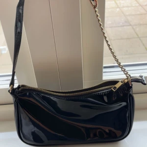Svart lackad axelväska med kedja - Snygg svart axelväska i glansigt lackmaterial med guldfärgad dragkedja och kedjedetalj på axelremmen. Enkel att matcha till olika outfits.
